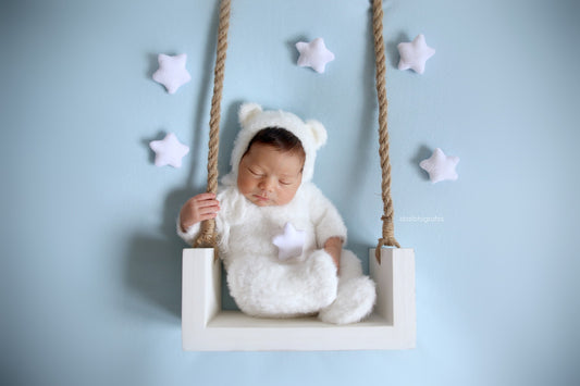 Newborn asleep on a white rustic swing, wearing a knit prop outfit. 