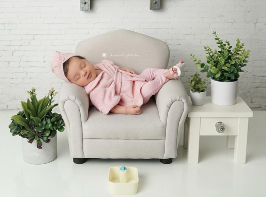  Newborn photography prop featuring a tiny beige armchair with a sleeping baby in a pink robe and leggings, surrounded by potted greenery and a small white side table, creating a cozy spa-inspired setup.