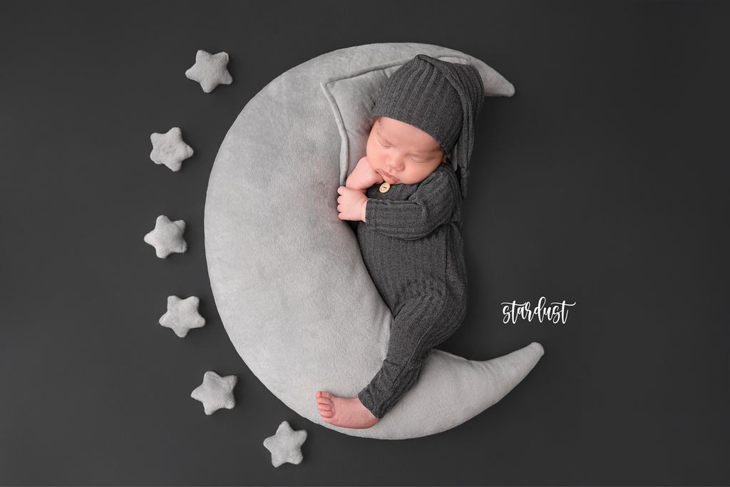 Newborn photography prop featuring a baby in a grey knit outfit, sleeping on a crescent moon pillow surrounded by small star pillows on a dark background.