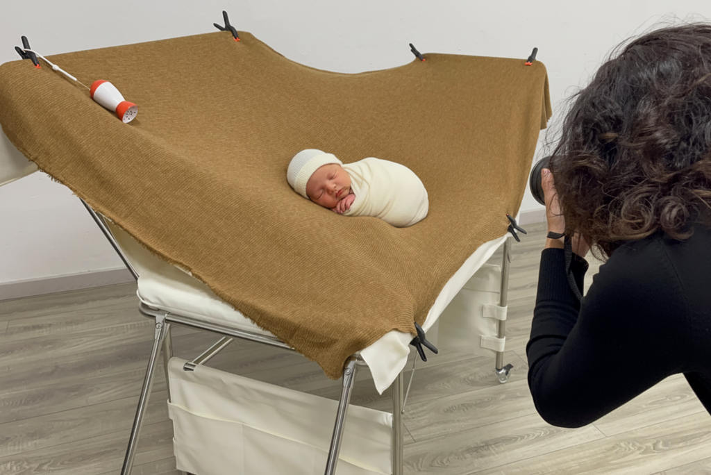 A baby photographer captures an image of a baby doll posed on an extended backdrop surface of a Newborn Photography Posing Table. The table is covered with a brown fabric that is secured with clips, showcasing the ample space for various posing options. The setup highlights the functionality and stability of the posing table for professional newborn photography.