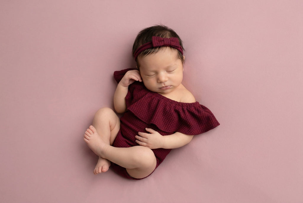 Newborn baby in a burgundy outfit and headband on a pink background