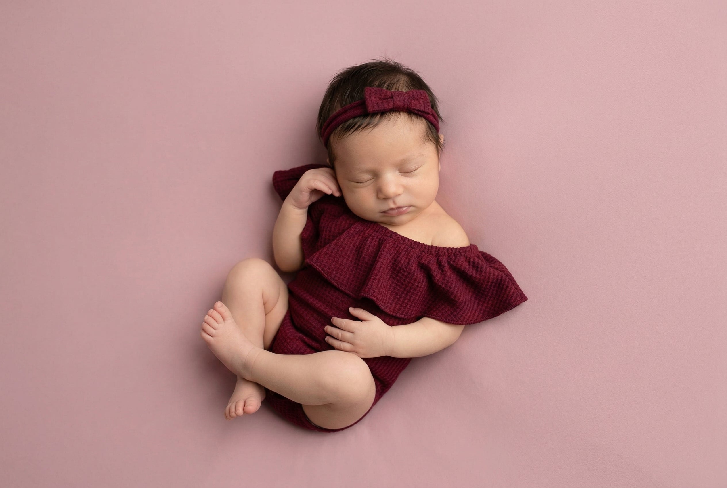 Newborn baby in a burgundy outfit and headband on a pink background