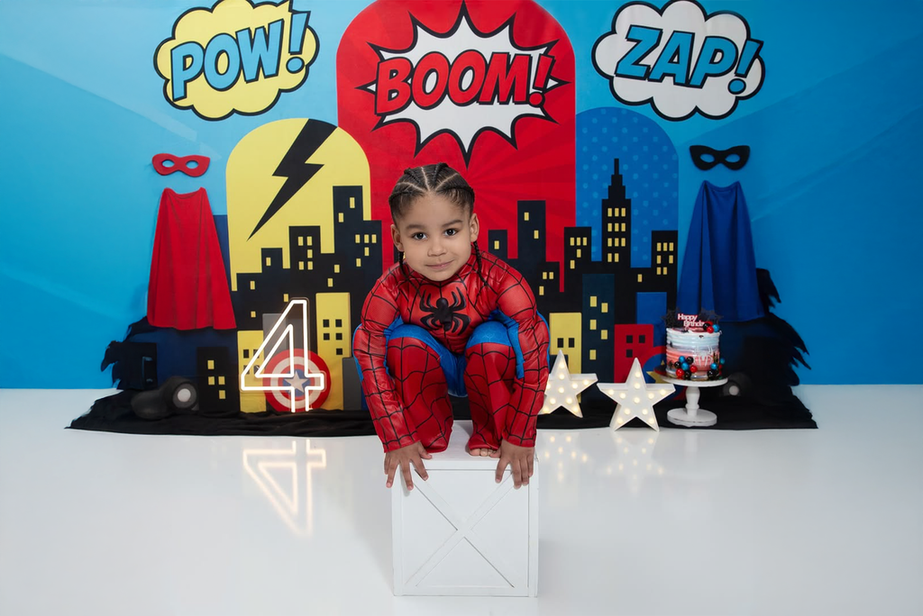 Child in superhero costume with comic-themed backdrop on top of cake smash box prop