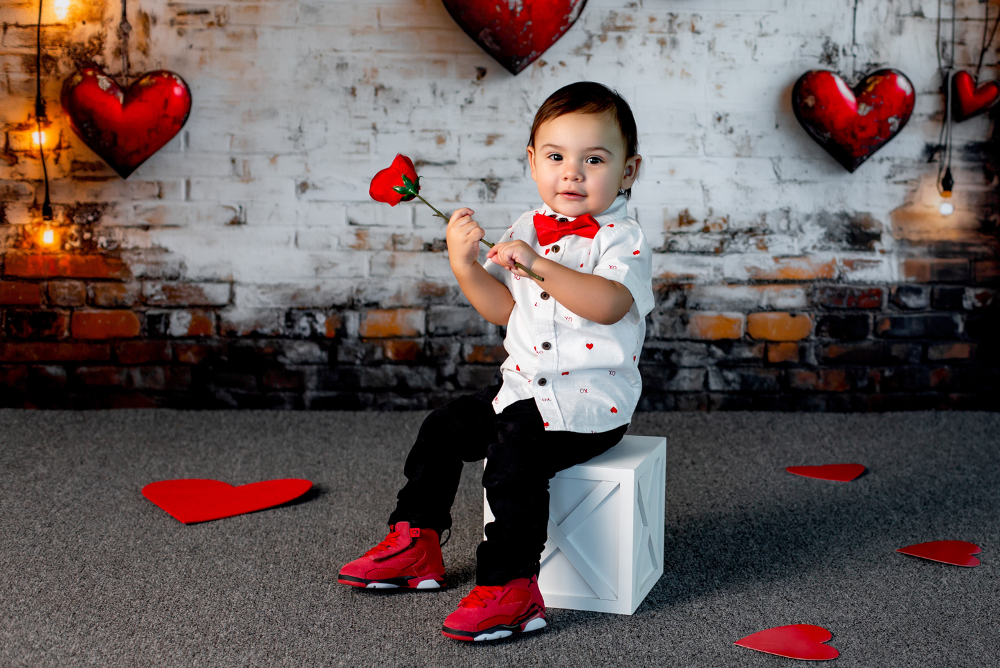 A toddler sits on a white farmhouse X-paneled cake smash box, holding a red rose in a Valentine-themed photoshoot. The backdrop features a rustic brick wall with glowing string lights and hanging red heart decorations. Red paper hearts are scattered on the floor, enhancing the romantic setting. This versatile newborn photography prop adds charm and elegance to themed sessions.