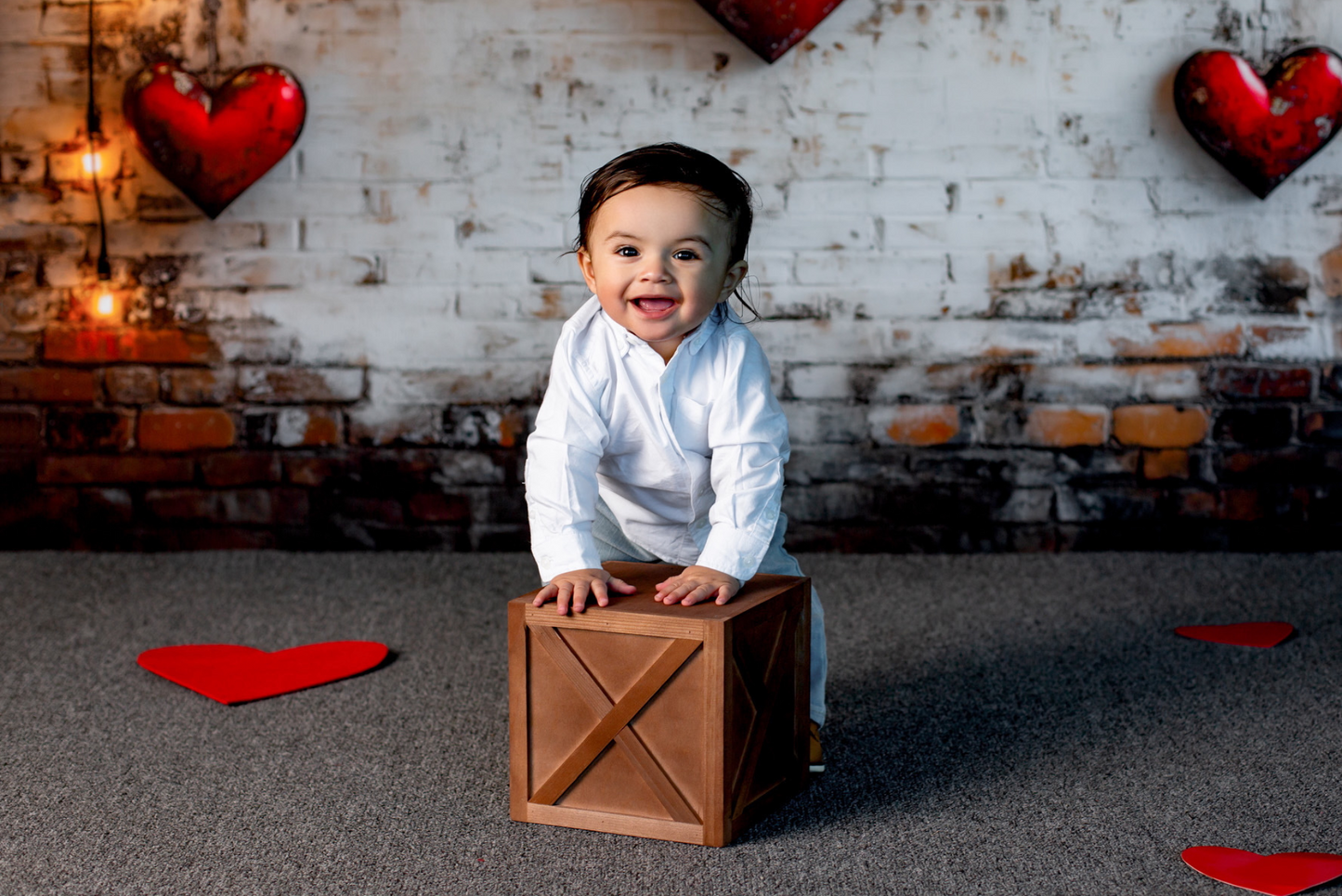 Smiling baby in a white outfit leans on a brown farmhouse X-paneled newborn photography prop box. The rustic brick wall, glowing string lights, and red heart decorations create a cozy Valentine’s-themed setup.