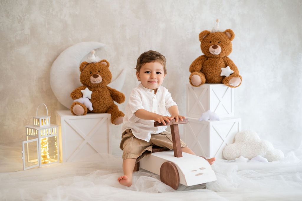 Smiling toddler on wooden ride-on surrounded by white cake smash boxes, a dreamy milestone photography prop.