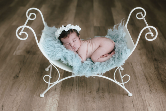 Newborn photography prop: A peaceful baby sleeps on a white vintage curved bench, adorned with a blue fluffy blanket and a white flower headband, set against a wooden floor backdrop.