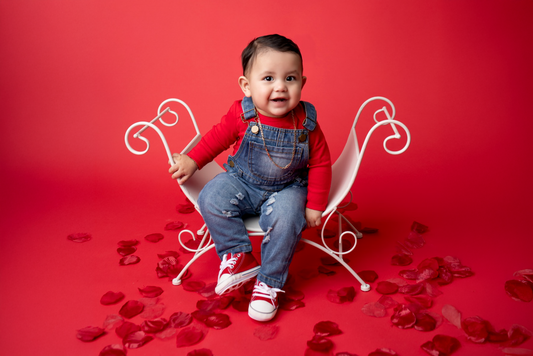 Smiling baby in denim overalls and red sneakers sits on a curved vintage bench, a charming newborn photography prop. The red backdrop and scattered rose petals create a warm, romantic setting.