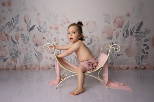 A baby sits on a curved vintage bench, painted light yellow with a soft pink wrap, against a floral backdrop. The scene features a newborn photography prop, creating a delicate, pastel-toned setting.