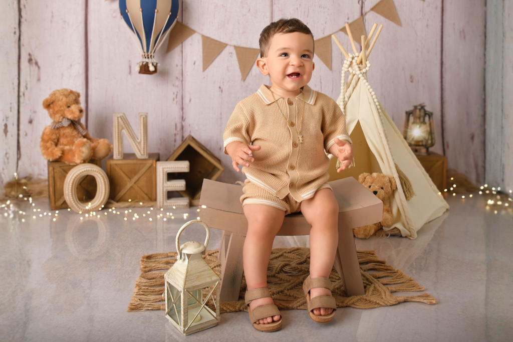 Child in a beige outfit sitting on a stool curved bench with teddy bears and decorative items for milestone photo session.