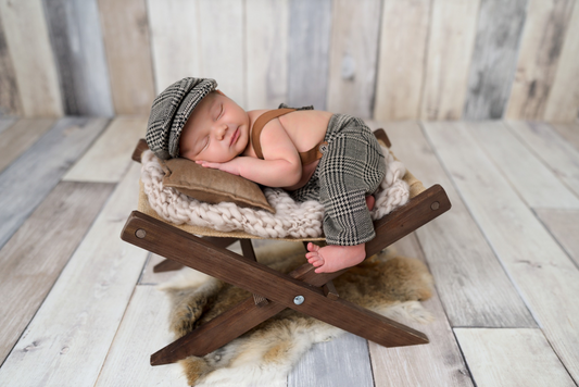 Newborn photography prop: Baby in houndstooth cap and suspenders sleeps on a rustic wooden chair with cozy knit blankets and a soft brown pillow.