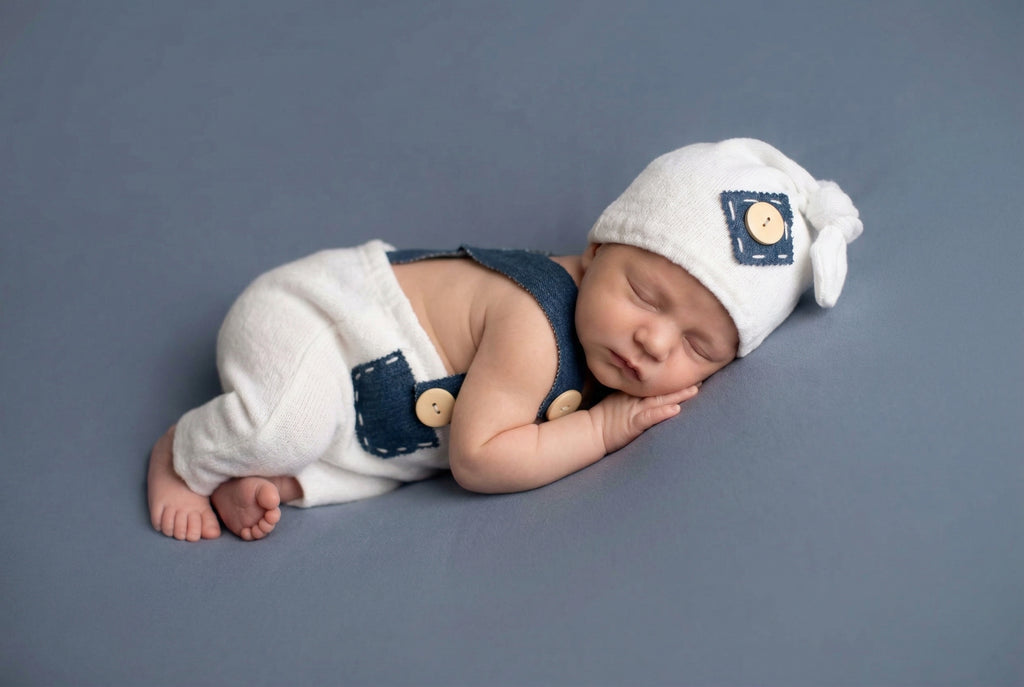 Newborn baby wearing a white hat and navy overalls on a gray background