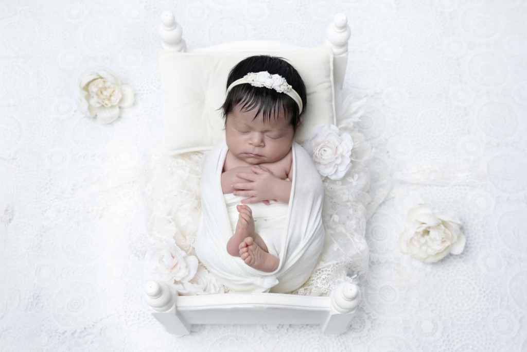 Wrapped newborn posed in white mini bed with lace and flowers, elegant newborn photography prop.