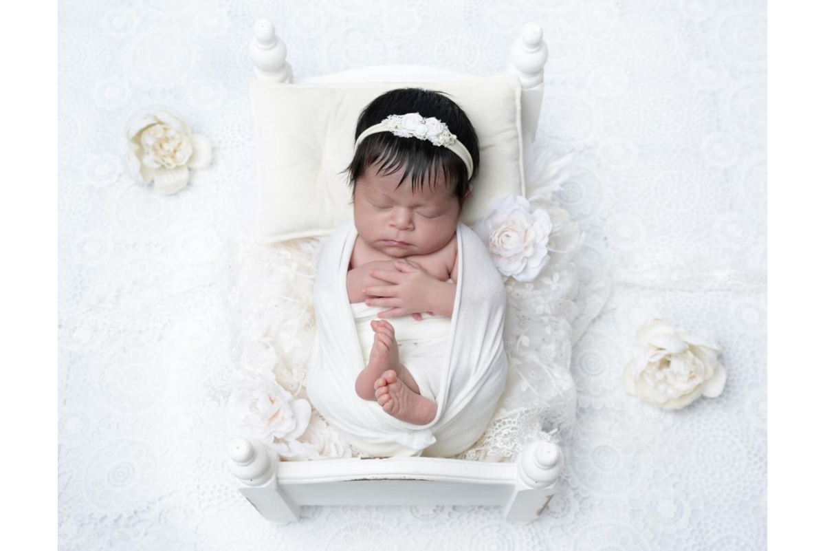 Newborn peacefully sleeping in a small white wooden bed, swaddled in a white wrap with a floral headband. The setup includes lace fabric and white flowers, emphasizing the delicate newborn photography prop.