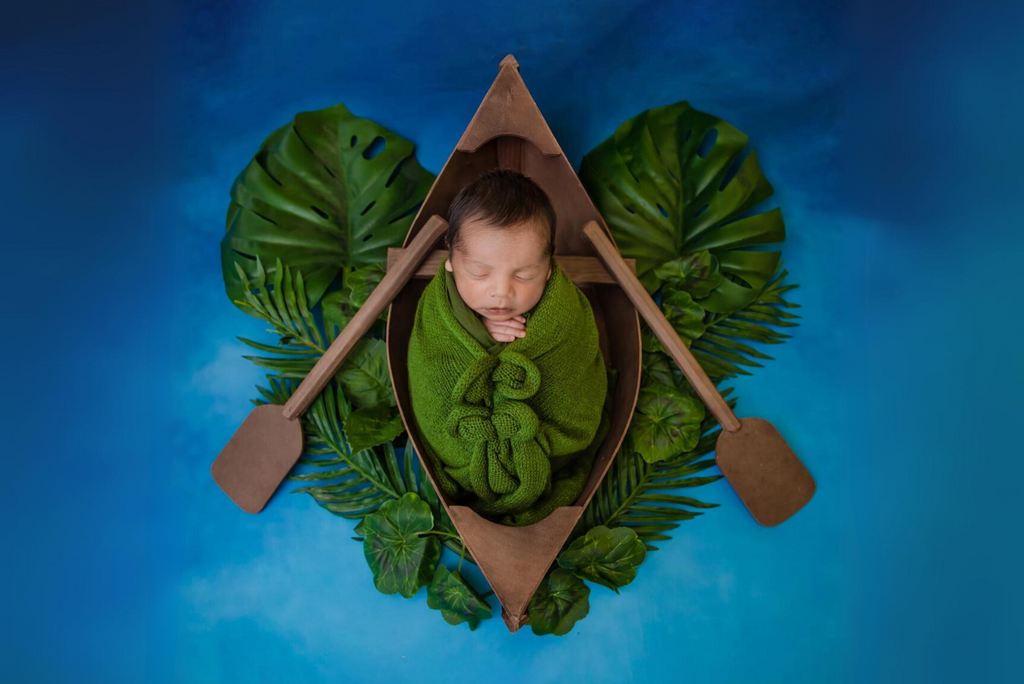 Newborn wrapped in green blanket, sleeping in a wooden canoe newborn photography prop with tropical leaves.
