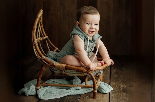 Smiling baby sitting on a half-moon rattan hammock chair, a beautiful newborn photography prop. The rustic wood backdrop and soft sage-green wrap create a cozy, vintage-inspired setting.