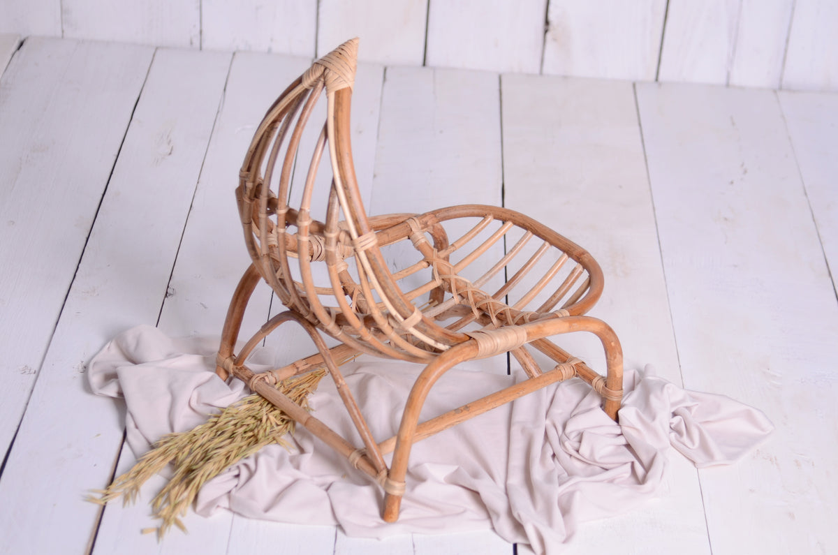 Handwoven rattan newborn photography prop in a crescent moon shape, placed on a whitewashed wooden floor with neutral fabric and wheat accents for a cozy, boho-inspired look.