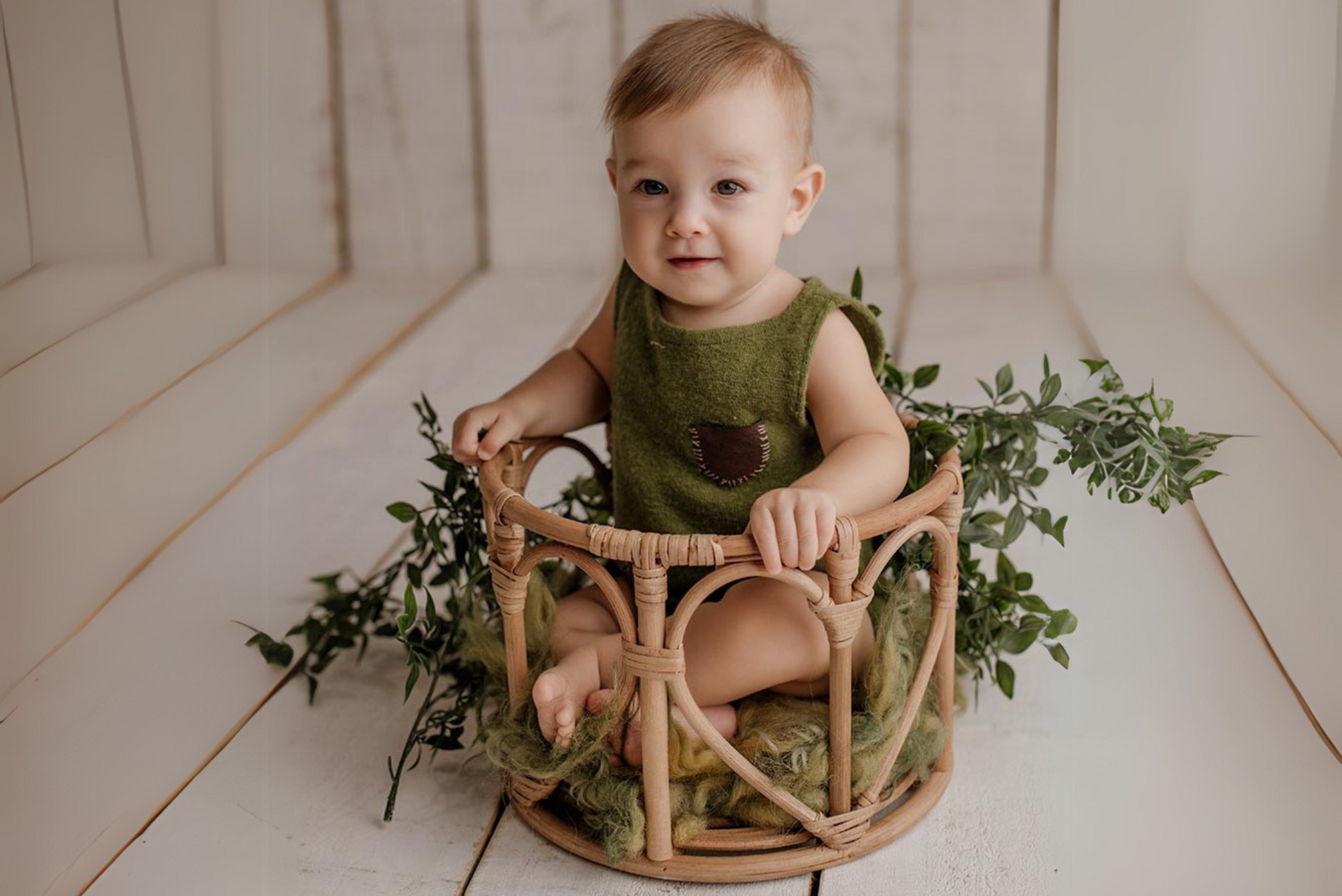 A baby in a green romper sits in a LovePod rattan basket, surrounded by greenery—perfect for newborn photography prop styling.