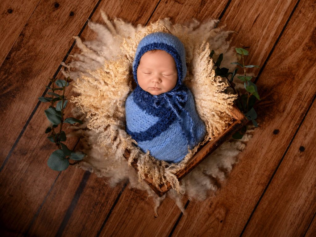 Newborn baby wrapped in blue fabric and bonnet, lying on a wooden floor with decorative elements.