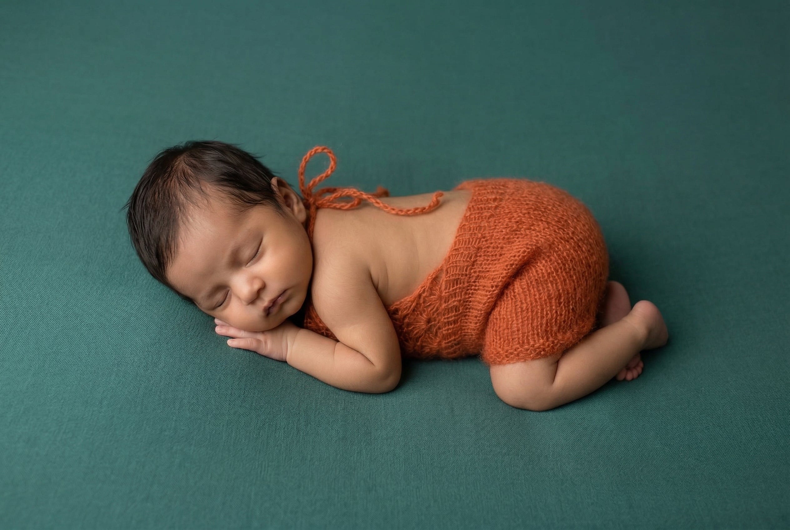 Newborn baby in orange knitted outfit lying on a teal background