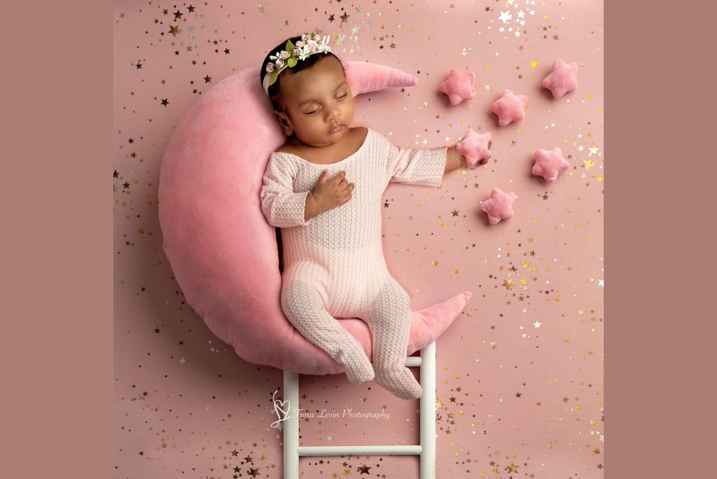 Baby in a pink outfit lying on a pink crescent moon-shaped pillow with star decorations.
