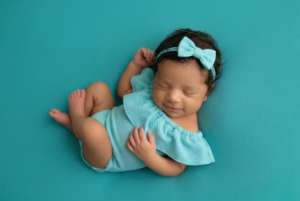 Newborn baby in a turquoise outfit with a matching headband on a teal background