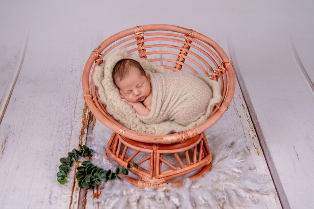 A sleeping newborn wrapped in a cream knit is curled in a boho papasan rattan chair newborn photography prop.