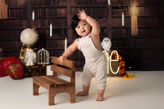 Adorable baby in a white romper stands beside a wooden bench in a cozy library-themed setup, featuring a magical globe and owl decor. Newborn photography prop.