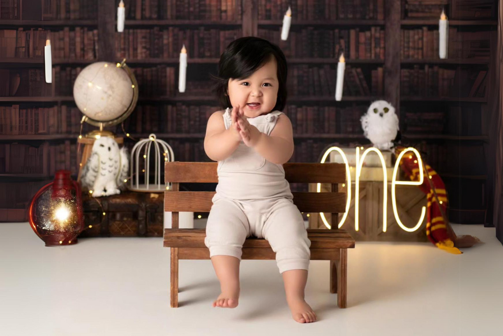 Smiling baby in a white romper sits on a wooden bench, clapping hands in a charming library-themed setting with owl and globe props. Newborn photography prop.
