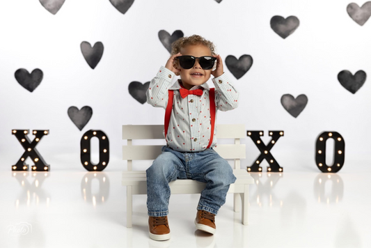 A cheerful toddler sits on a small white bench, wearing sunglasses, a red bow tie, and suspenders, with a Valentine's Day-themed backdrop featuring black heart decorations and illuminated "X" and "O" letters