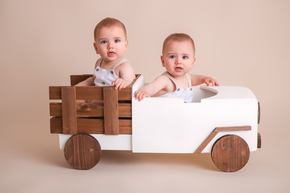 Twin babies sit in a wooden and white convertible pickup truck, a charming newborn photography prop.