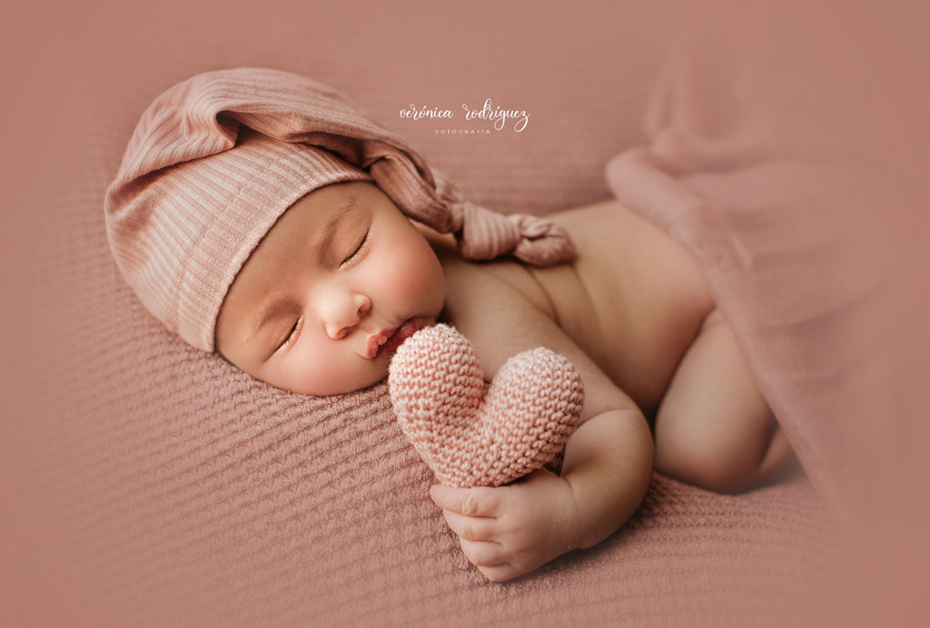 Newborn baby holding a pink knitted heart on a soft pink background wearing a Pink ribbed beanie.