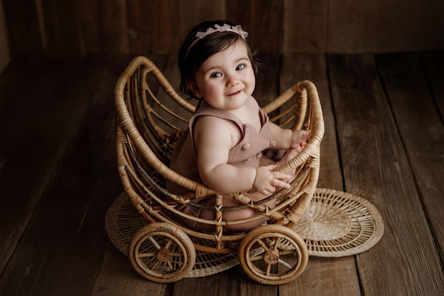 Smiling baby girl sits inside a rattan baby carriage, a charming newborn photography prop, on rustic wooden flooring. She wears a soft pink romper and matching headband, creating a warm, vintage-inspired scene.