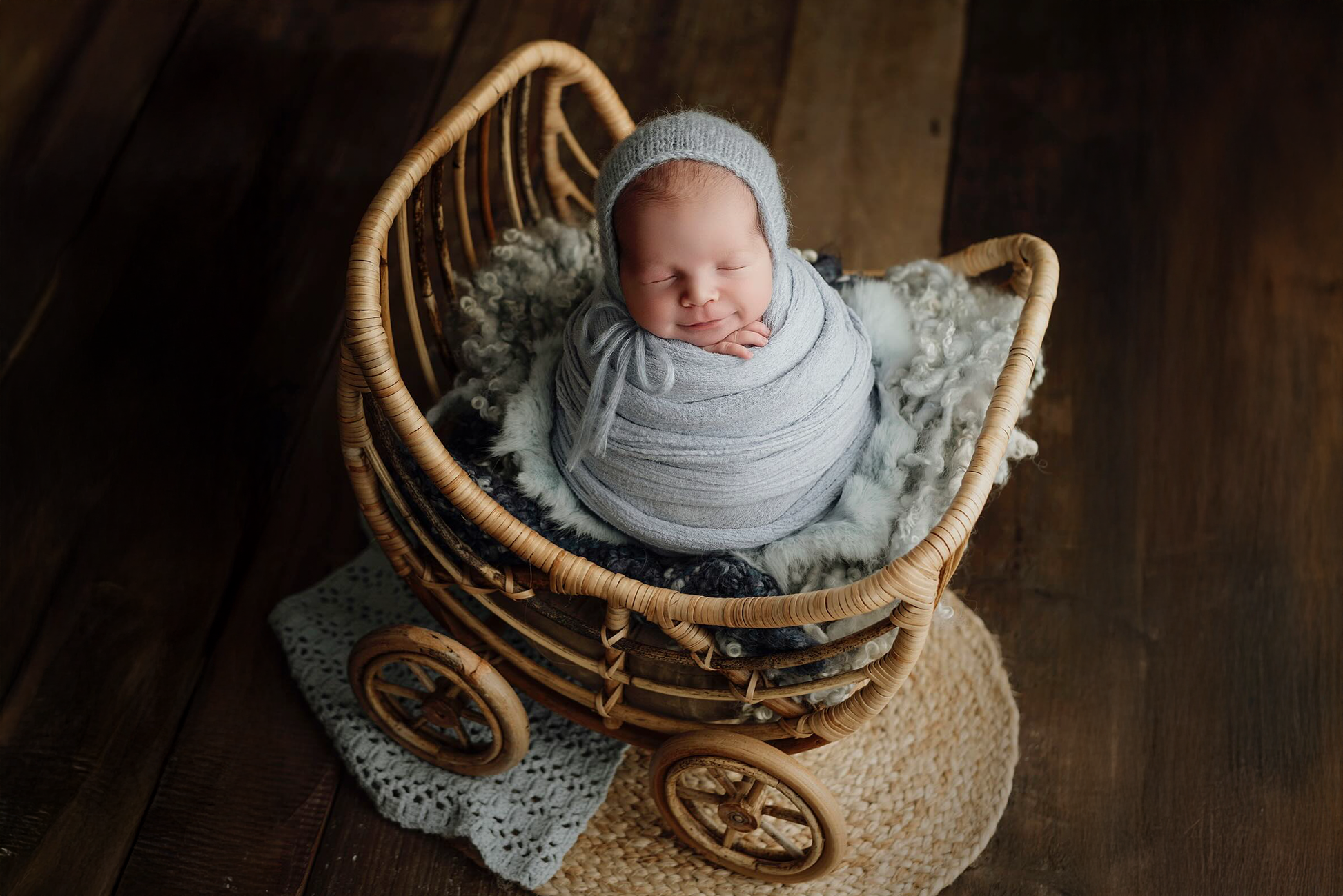 Newborn photography prop: A vintage-style rattan baby carriage holds a peacefully swaddled baby in soft gray wraps, resting on plush wool layers. A dreamy, rustic setup on a wooden floor.