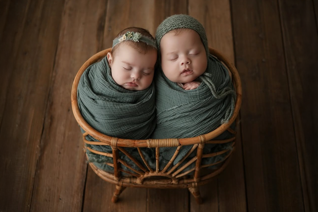 Adorable newborn twins peacefully sleeping in the Sun-Kissed Rattan Cradle prop, wrapped in soft sage green swaddles. One baby wears a delicate floral headband, while the other is adorned with a matching knit cap. The rustic wooden floor complements the natural, handwoven rattan design of the cradle, perfect for capturing precious newborn moments. Ideal for eco-conscious photographers seeking a stylish and safe baby prop for their photography studio.