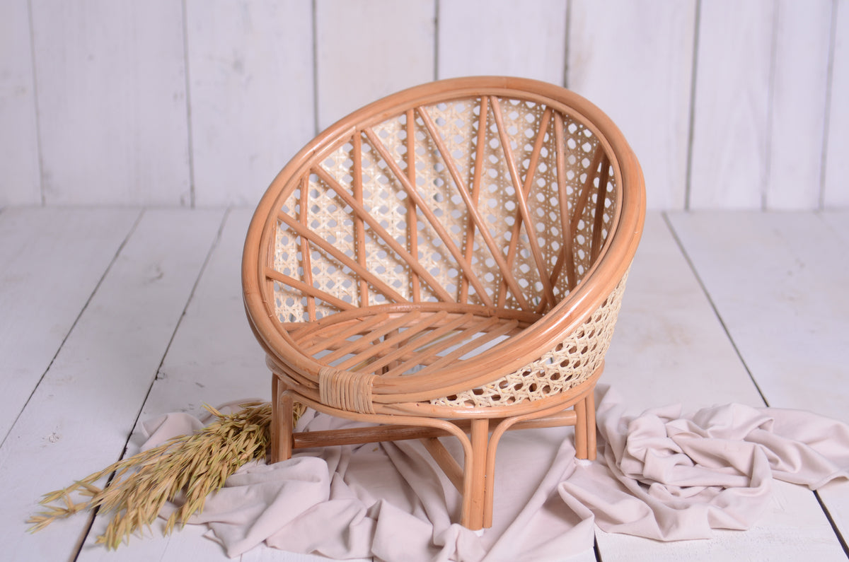 Elegant rattan nest cocoon newborn photography prop with intricate cane weaving, set on a rustic white wood backdrop with soft beige fabric and dried wheat.