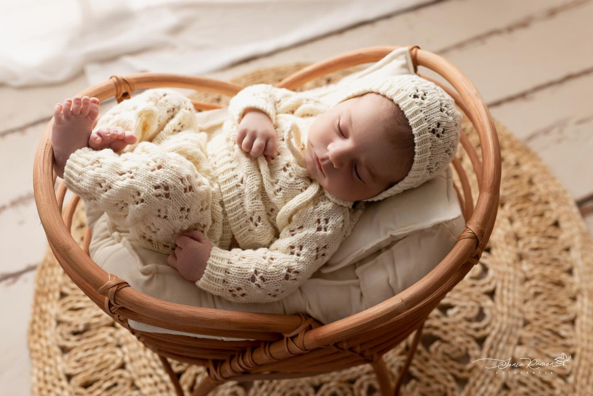 Newborn nestled in a cozy rattan cradle, dressed in a cream knit onesie and bonnet. Newborn photography prop.