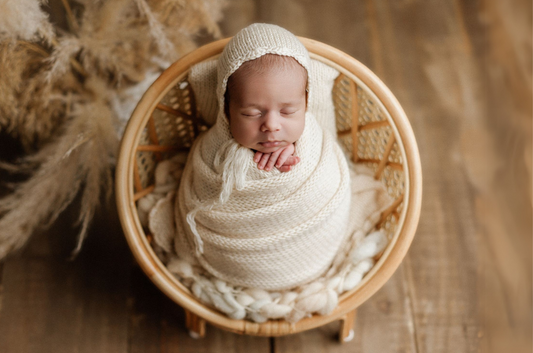 Newborn photography prop featuring a rattan nest chair with a swaddled baby in a cozy cream knit wrap and bonnet. The soft wool layer adds warmth, while pampas grass and wood tones create a rustic charm.