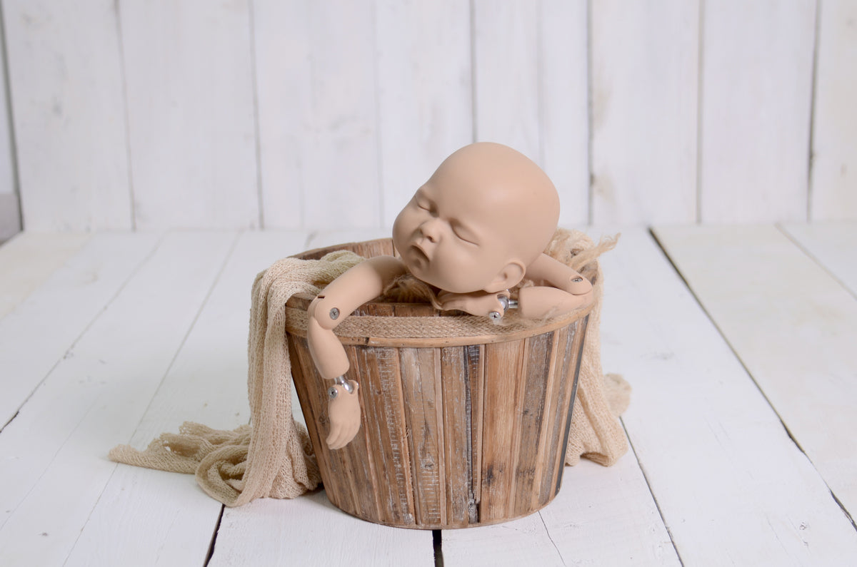 A newborn photography prop setup showcasing a newborn baby doll resting in a rustic wooden bucket, accented with a soft beige wrap. The scene is set on a white wood floor, creating a clean and airy aesthetic ideal for bright, timeless newborn portraits.