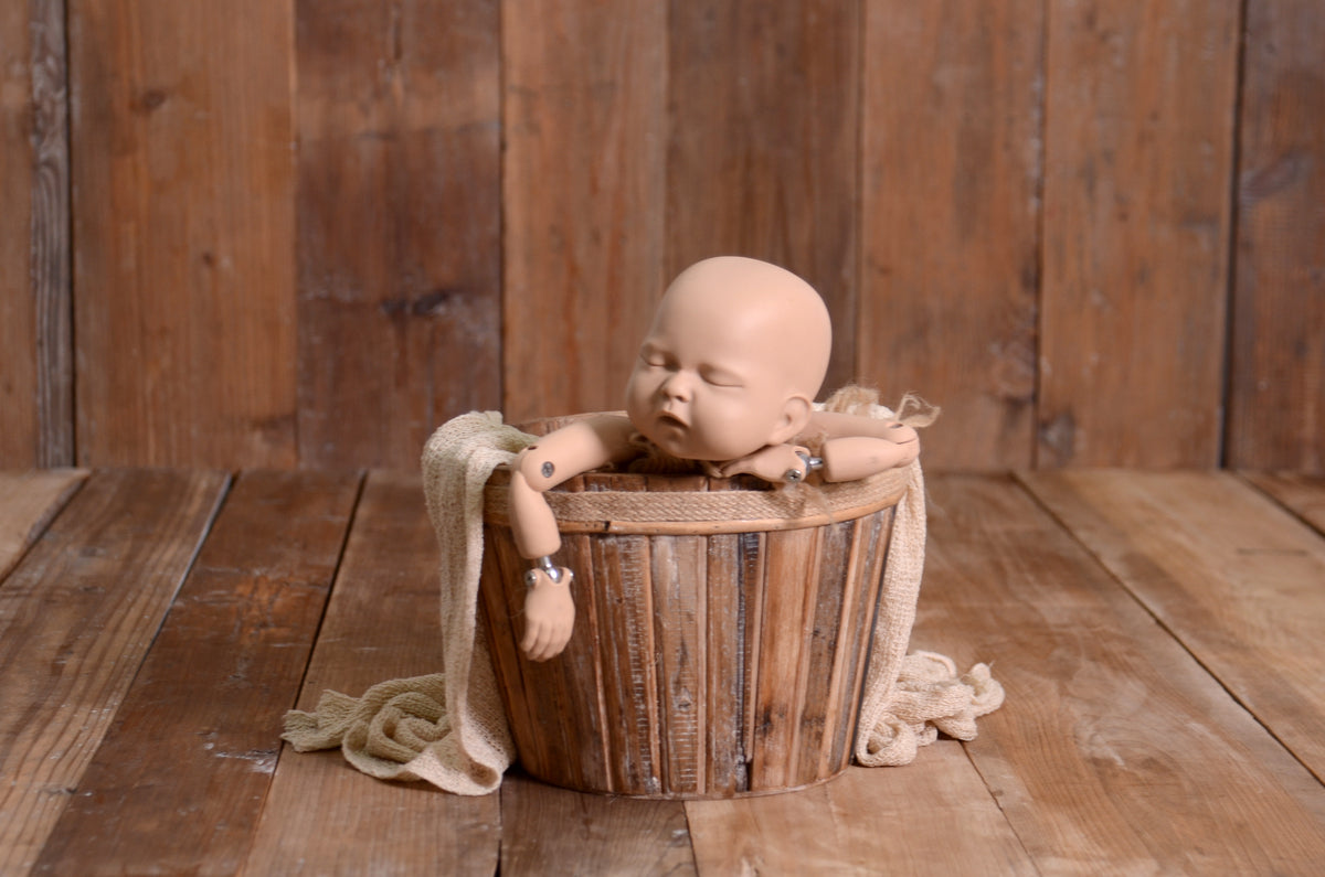 A newborn photography prop setup featuring a realistic baby posed in a rustic wooden bucket. The bucket is adorned with a soft beige wrap, placed against a brown weathered wood floor and matching backdrop for a warm, natural look. Perfect for creating cozy, timeless newborn portraits.