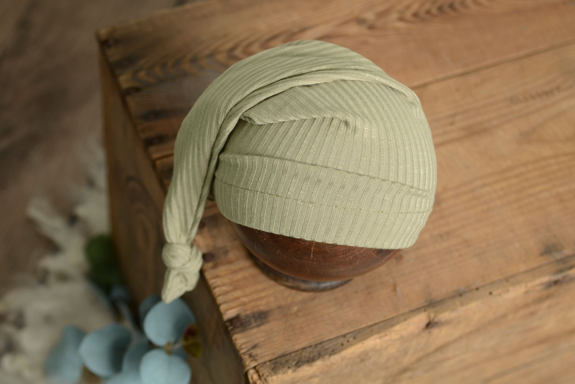 Sage green ribbed knot sleepy cap newborn photography prop styled on wooden bowl with soft rustic tones.