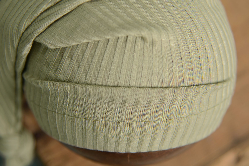 Sage green ribbed knot sleepy cap newborn photography prop close-up on rustic wooden bowl.