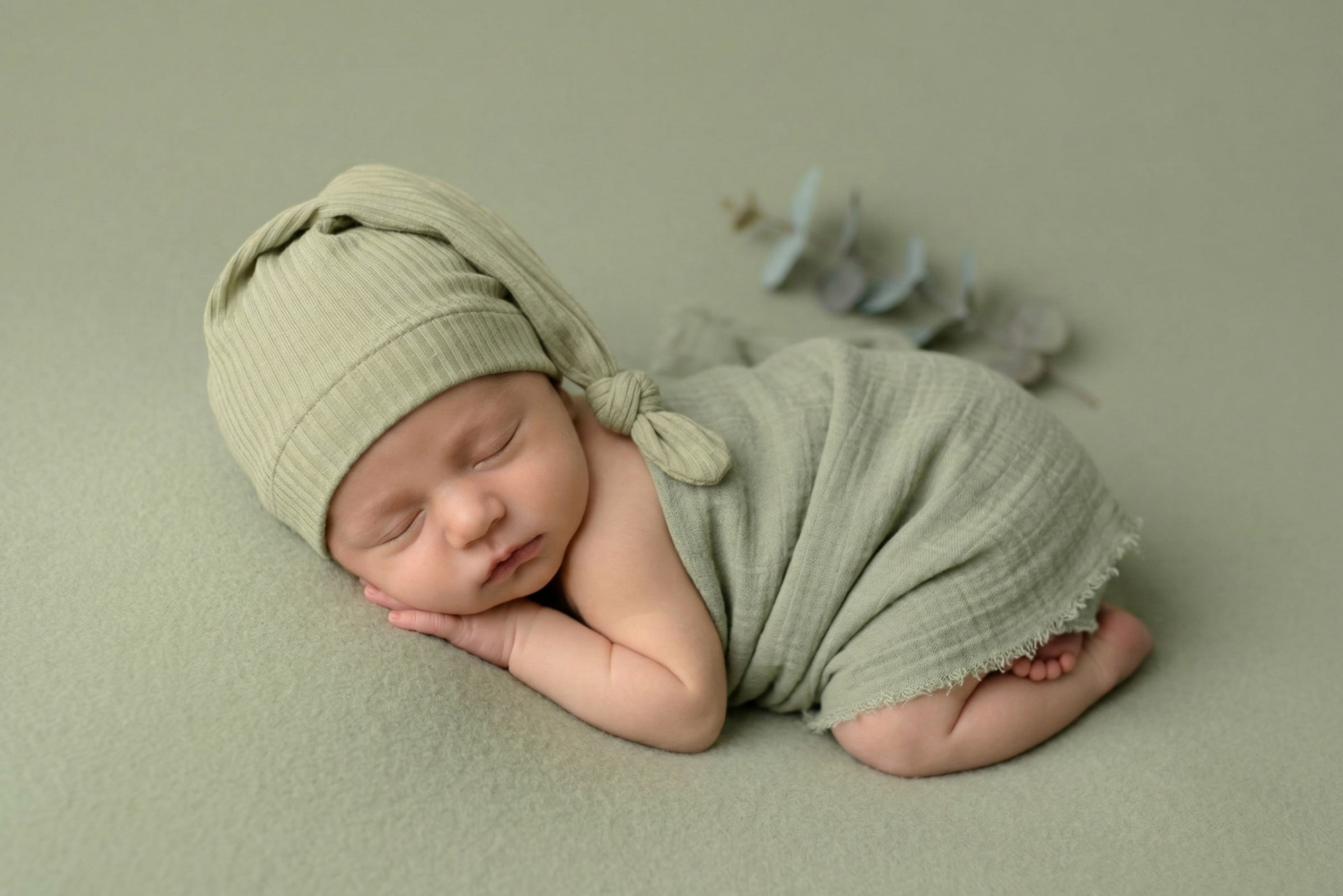 Sleeping newborn in sage wrap with ribbed knot sleepy cap newborn photography prop, soft green setup.