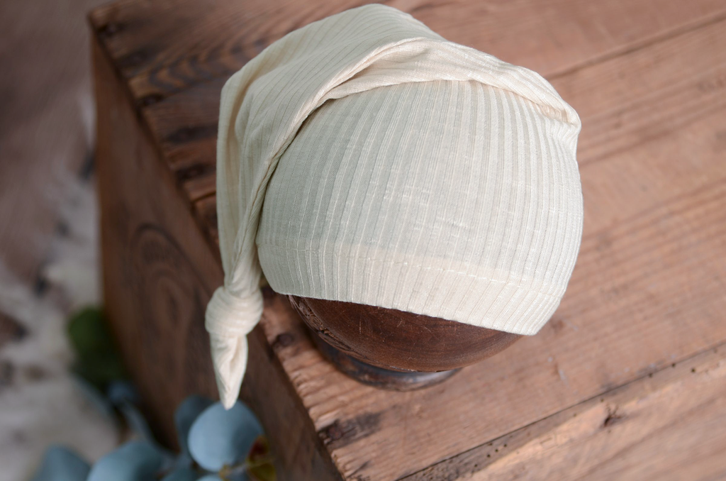 Cream ribbed newborn photography prop sleepy hat styled on a rustic wooden stand with soft blue florals.