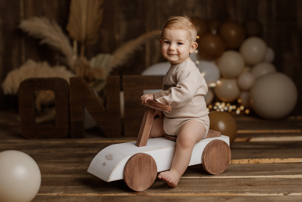 Toddler in a rustic outfit sits beside the Rustic Ride-On - White in a cozy, dark wood setting, creating a warm and vintage newborn photography scene.