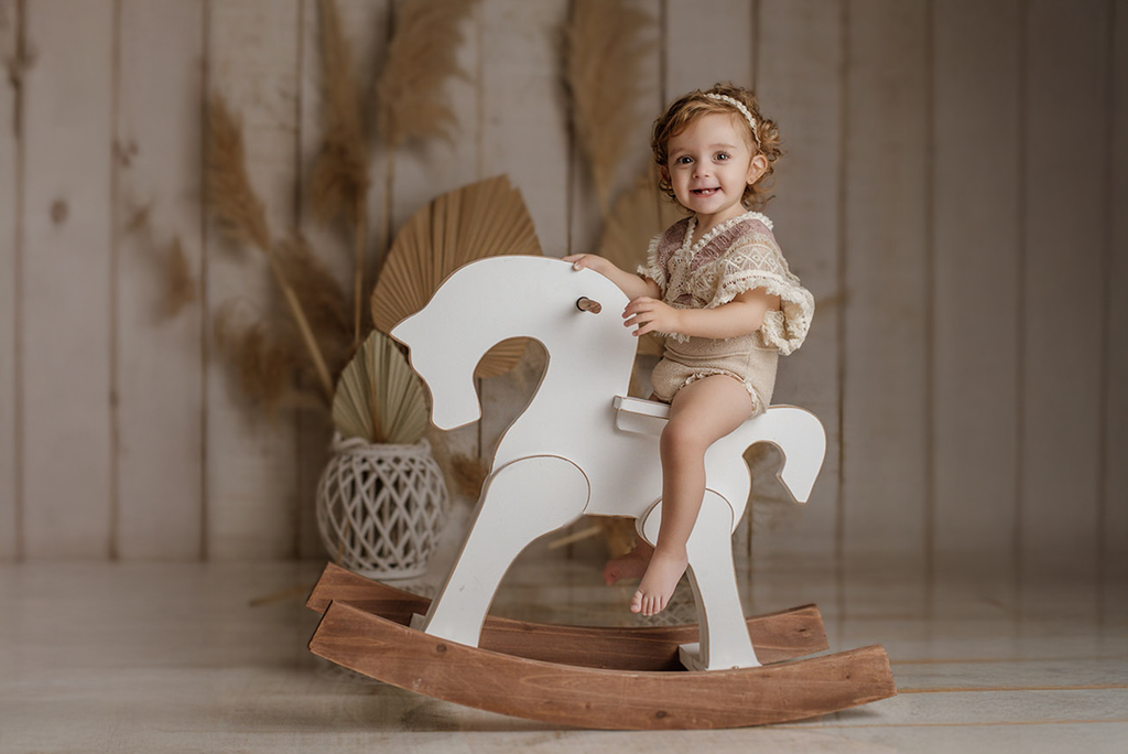 Toddler sits on a white wooden rocking horse, a charming newborn photography prop, in a neutral boho setting.
