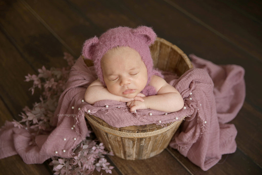 Newborn photography prop featuring a driftwood rustic bucket, adorned with a soft mauve wrap and pearl accents, cradling a baby in a cozy knitted bear bonnet.