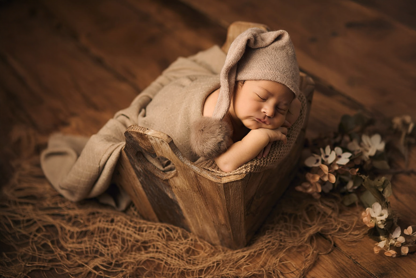 Newborn photography prop: a rustic wooden crate holds a peacefully sleeping baby wrapped in a soft beige swaddle with a matching bunny-ear hat. The cozy setup is styled with textured burlap and delicate floral accents, creating a warm, earthy aesthetic.