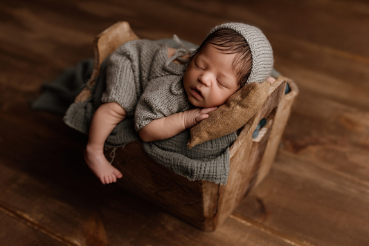 Newborn photography prop featuring a rustic wooden crate, with a baby in a cozy knit outfit and bonnet, peacefully sleeping on a soft pillow and textured blanket. Warm, earthy tones create a serene and timeless feel.