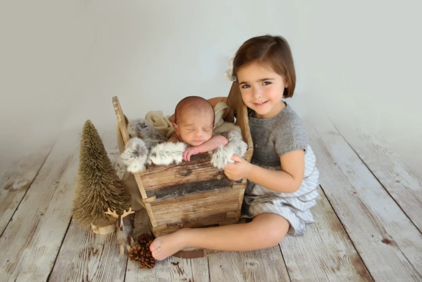 A newborn sleeps peacefully in a rustic wooden crate adorned with faux fur, accompanied by a smiling older sibling in a gray knit dress. The scene features a cozy newborn photography prop setup with a pinecone accent and a miniature tree, creating a warm, woodland-inspired atmosphere.